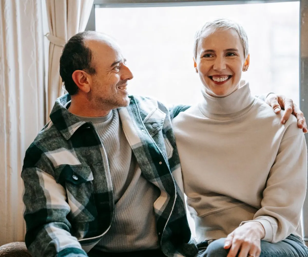 A smiling couple sitting close together beside a window.
